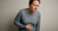 Middle-aged Caucasian woman experiencing stomach pain holding abdomen in gray sweater against neutral background for health and wellness concepts.