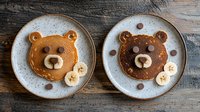 Cute bear-shaped pancakes decorated with chocolate chips and banana slices on rustic ceramic plates, creative breakfast food art on wooden table background.