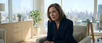 Middle-aged woman with brown hair and fair complexion, wearing dark business blazer, seated in modern office with city skyline view, professional corporate environment for business and leadership concepts.