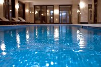 Indoor swimming pool with crystal clear blue water reflecting ambient lighting, surrounded by loungers and glass walls in luxury spa or hotel setting.