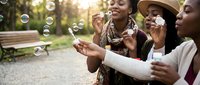 Young African American women friends enjoying soap bubbles together in sunny park setting creating joyful summer memories and carefree moments outdoors.