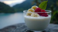 Fresh yogurt dessert with sliced banana and raspberry garnished with mint leaf against blurred mountain lake background, soft morning light.