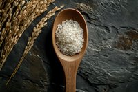 Raw white rice grains in wooden spoon on dark stone background with wheat ears, top view. Natural organic food ingredients for cooking.
