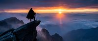 Silhouetted hiker standing on rocky mountain peak at sunrise above clouds, holding trekking poles, overlooking misty valley landscape with golden sun on horizon.