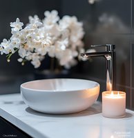 Modern bathroom interior with white vessel sink, chrome faucet, lit candle and delicate white orchids on marble countertop against dark wall, creating spa-like atmosphere.