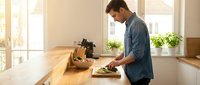 Young Caucasian man in denim shirt chopping vegetables on wooden cutting board in bright modern kitchen with natural sunlight streaming through windows.