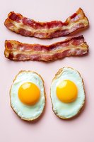 Crispy bacon strips and sunny side up fried eggs on pink background. Classic breakfast combination for morning meal concepts and culinary designs.