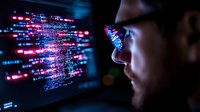 Young Asian male programmer analyzing code on screen with glowing blue and pink digital data reflection in glasses, working in dark environment.