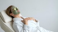 Young Caucasian woman relaxing in white bathrobe with green clay face mask and cucumber slices on eyes at spa salon.