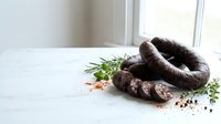 Artisanal dark blood sausage with fresh herbs and spices on white marble surface near window for gourmet cooking and traditional cuisine recipes.