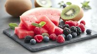 Fresh watermelon slices with mixed berries, kiwi, and mint leaves arranged on dark slate board, healthy summer fruit composition for refreshing snacks.