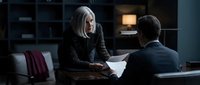 Senior woman with long gray hair and middle-aged man in dark clothing reviewing documents together at desk in modern office setting with shelving and warm lighting.