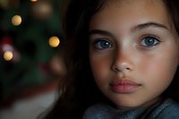 Young girl with blue eyes and dark hair gazes directly at camera with serious expression, warm bokeh lights in soft-focus background suggesting holiday setting.