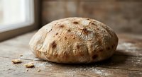 Freshly baked artisan bread loaf with golden crust and flour dusting on rustic wooden surface near window for bakery and homemade cooking concepts.