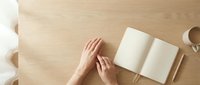 Hands reaching for open blank notebook on wooden desk with coffee cup, top view workspace setup for writing, planning and creative work.