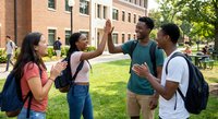 Diverse college students celebrating success with high five on university campus lawn surrounded by brick dormitory buildings.