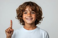 Young child with curly brown hair wearing white t-shirt showing index finger up with bright cheerful smile against light gray background, studio portrait.
