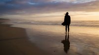 Silhouetted surfer holding board standing on wet sand beach at golden hour sunset, contemplating ocean waves, solitude and coastal lifestyle.