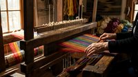 Elderly woman weaving colorful striped textile on traditional wooden loom in rustic workshop with natural lighting creating authentic handcraft atmosphere.