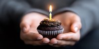 Hands holding chocolate cupcake with single burning birthday candle against dark background, intimate celebration moment captured in panoramic format.