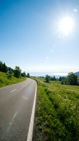 Curved asphalt road through lush green valley with forested hills, bright sunlight, and distant mountains under clear blue sky. Perfect for travel, transportation, and scenic landscape projects.