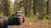 Hand in knitted glove holding stainless steel thermos mug in autumn forest setting for outdoor adventure and camping activities.