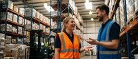 Warehouse workers discussing logistics in modern distribution center with shelving and forklift equipment for supply chain management operations.