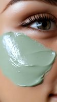 Close-up of green clay facial mask on young woman's cheek with focused view of eye with long eyelashes and hazel iris, showing skincare treatment application.