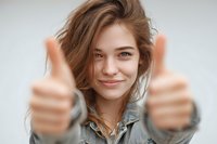Happy young woman with brown hair giving thumbs up gesture showing positive attitude and success in casual denim shirt against neutral background.