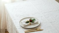 Minimalist table setting with white plate, gold cutlery, and fresh herb garnish on textured cream tablecloth for fine dining and hospitality design projects.