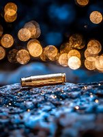 Empty bullet casing on wet surface with golden bokeh lights in background creating dramatic atmosphere. Artistic macro photography with blue tones.