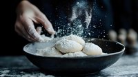 Dusting powdered sugar over freshly baked cookies in dark ceramic plate, dramatic moody lighting with flour particles floating in air against black background.
