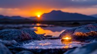 Dramatic sunset over mountain lake with sunlight reflecting off wet rocks in foreground, creating golden highlights against dark silhouettes and moody purple clouds.