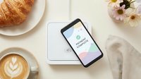 Smartphone displaying wellness app interface on white tablet surrounded by croissant, latte coffee with latte art, fresh flowers and napkin for lifestyle and technology marketing.