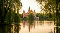 Romantic castle reflected in calm lake waters at golden hour sunset, framed by weeping willow trees creating peaceful fairy tale landscape for travel content.