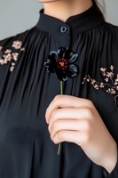 Female hand holding single black cosmos flower against pleated black dress with mandarin collar and delicate pink floral embroidery details. Elegant fashion styling.