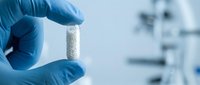 Scientist in blue latex gloves holding a white cylindrical pharmaceutical tablet or capsule in laboratory setting with blurred medical equipment background.