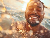 Young African American man laughing with joy while swimming in water at sunset, with water droplets and golden bokeh light creating magical summer atmosphere.