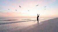 Person celebrating freedom with victory gesture on peaceful beach at sunset with flying birds creating inspirational moment for wellness and motivation content.