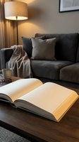 Open blank book on wooden coffee table in cozy living room with gray sofa, warm lamp light, mug, and glasses in background.