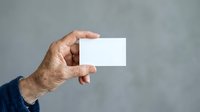 Senior man holding blank white business card against neutral gray background for corporate branding and professional identity mockup.