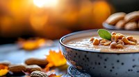 Creamy pumpkin soup garnished with roasted pine nuts and fresh herbs in black ceramic bowl, autumn leaves and warm bokeh background creates cozy atmosphere.