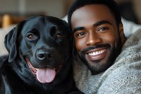 Young African American man with bright smile embracing black Labrador retriever dog, close-up portrait showing bond between pet owner and companion animal.