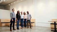 Diverse business team of five professionals standing together in modern minimalist office space with white brick wall and wooden furniture.