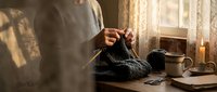 Woman knitting dark wool sweater at cozy home workspace with warm lighting, crafting handmade clothing in comfortable domestic setting for relaxation.