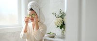 Woman in white bathrobe and towel holding cucumber slices over eyes for natural skincare routine in bright bathroom spa setting.