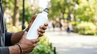 Man with dark skin tone holding blank white bottle outdoors in park setting, suitable for beverage, health, fitness, and eco-friendly product marketing campaigns.