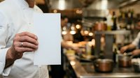 Man in white chef's coat holding blank white paper in professional kitchen environment with blurred background and warm lighting.