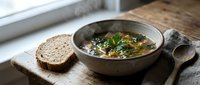 Hearty vegetable soup with fresh herbs served in rustic ceramic bowl alongside artisan bread slices on wooden cutting board for cozy meal preparation.