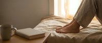 Woman relaxing on bed with book and coffee cup in warm morning sunlight streaming through window creating cozy bedroom atmosphere for rest and leisure.